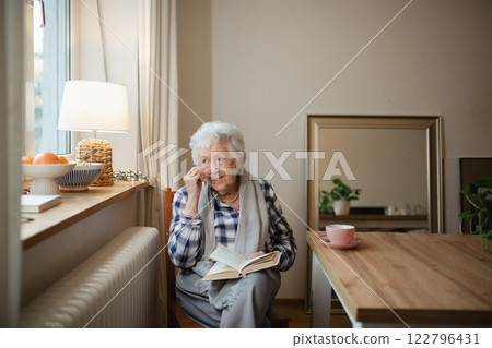 Lovely elderly lady reading a book, sitting at the kitchen table and sipping tea. Lovely elderly lady reading a book, sitting at the kitchen table and sipping tea. 122796431