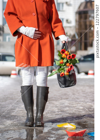 Closeup of woman in a orange coat and rubber boots walking down the street with a bouquet of tulips 122797607