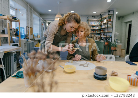 Happy retired student learns new skills, creating a handmade ceramic bowl. Female artist teaches an elderly woman how to shape clay at a hands-on master class Happy retired student learns new skills, creating a handmade ceramic bowl. Female artist teaches an elderly woman how to shape clay at a hands-on master class 122798248