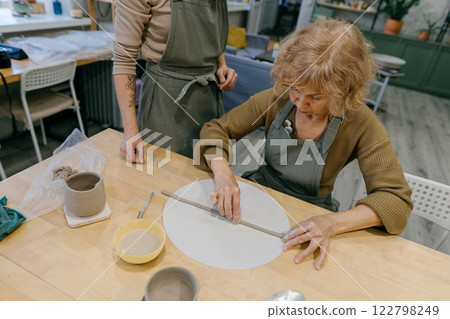 Teacher guides an elderly women, demonstrating sculpting techniques in a hands-on class. Elderly Caucasian woman learns pottery at a creative workshop with a young mentor Teacher guides an elderly women, demonstrating sculpting techniques in a hands-on class. Elderly Caucasian woman learns pottery at a creative workshop with a young mentor 122798249
