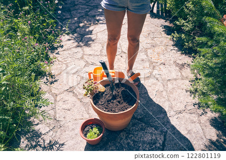 A woman's feet near a pot of earth. When transplanting plants, it is necessary to water the soil. The brunette is going to transplant plants into a clay pot. High quality photo 122801119