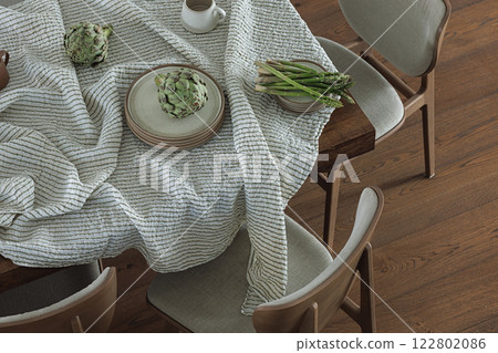 Simple organisation of dining table with ceramic dinnerware, linen tablecloth, dining chairs, artichoke and asparagus. Home decoration concept Simple organisation of dining table with ceramic dinnerware, linen tablecloth, dining chairs, artichoke and asparagus. Home decoration concept 122802086