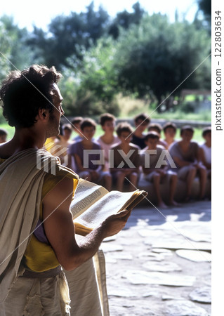 Aristotle teaches a group of teenagers using ancient papyrus in an outdoor setting 122803634