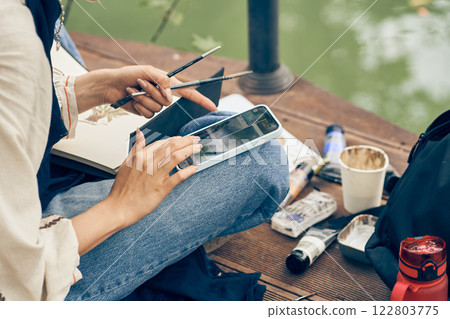The artist's hands are holding a smartphone, the girl is painting outdoors. Art supplies stand side by side against the background of a green lake. High quality photo 122803775
