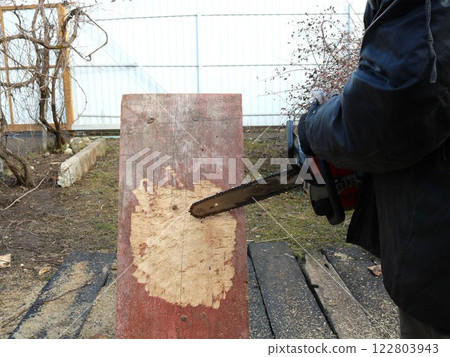 sawing out a part of a board with a chainsaw in a carpenter's shop outside, cutting off chips from a solid old board with a chainsaw blade to create a designer object outdoor sawing out a part of a board with a chainsaw in a carpenter's shop outside, cutting off chips from a solid old board with a chainsaw blade to create a designer object outdoor 122803943