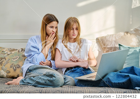 Two young serious teenage girls friends students at home looking at laptop, sitting on couch 122804432