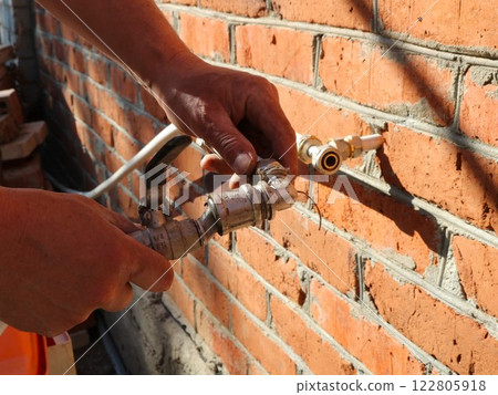 installation of street drainage system, hands holding a tap and a bent metal pipe to connect to a plastic pipe coming out of a brick wall installation of street drainage system, hands holding a tap and a bent metal pipe to connect to a plastic pipe coming out of a brick wall 122805918