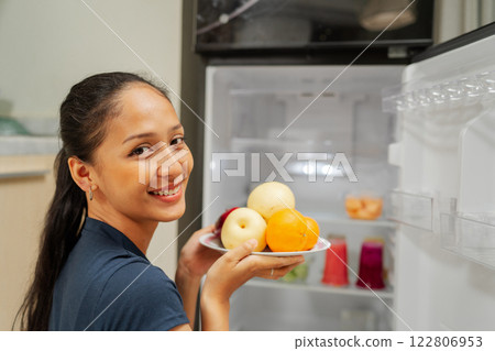 A Young Woman Holding Fresh and Colorful Fruits Near an Open Refrigerator Door A Young Woman Holding Fresh and Colorful Fruits Near an Open Refrigerator Door 122806953