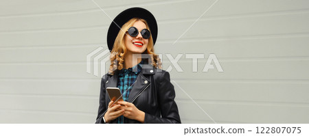 Stylish happy smiling modern young woman with phone, girl holding smartphone in black hat, jacket 122807075