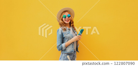 Summer modern happy young woman holding a smartphone in straw tourist hat on yellow wall background 122807090