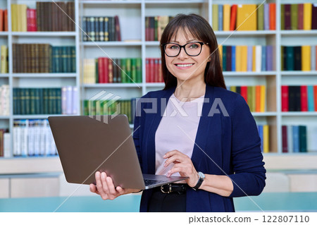 Portrait of middle aged smiling woman teacher professor with laptop inside library 122807110