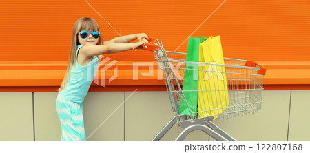 Happy little girl child walking with trolley cart and colorful shopping bags on city street 122807168