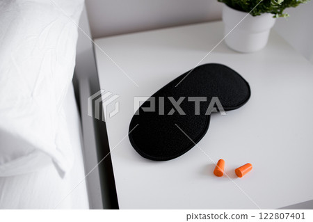 Black sleep mask and orange earplugs on white nightstand with green plant 122807401