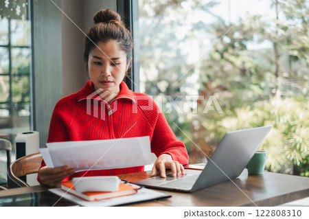 Woman in a red sweater working at a table with a laptop and documents, sitting by a window with greenery outside. Woman in a red sweater working at a table with a laptop and documents, sitting by a window with greenery outside. 122808310