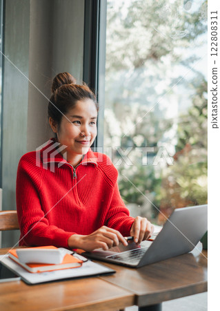 A young woman in a red sweater uses a laptop at a cafe, surrounded by natural light and greenery. A young woman in a red sweater uses a laptop at a cafe, surrounded by natural light and greenery. 122808311