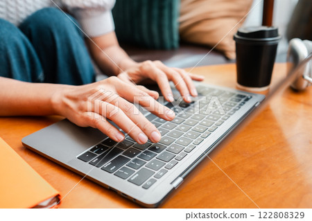 Hands typing on a laptop keyboard with a coffee cup nearby, set on a wooden table in a cozy indoor environment. Hands typing on a laptop keyboard with a coffee cup nearby, set on a wooden table in a cozy indoor environment. 122808329