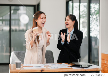 Two businesswomen in a modern office, laughing and celebrating success with documents and coffee on the table. Two businesswomen in a modern office, laughing and celebrating success with documents and coffee on the table. 122808338