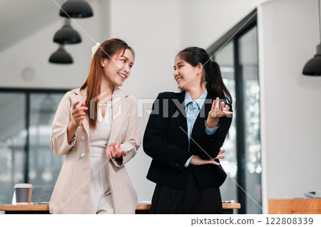 Two women in business attire share a light moment in a contemporary office environment, showcasing teamwork and collaboration. Two women in business attire share a light moment in a contemporary office environment, showcasing teamwork and collaboration. 122808339