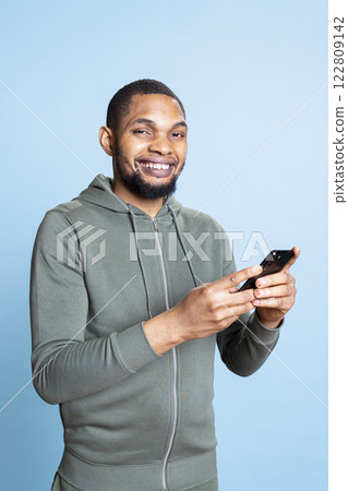 African american confident man browsing online websites and using social media, answering his texts and smiling on camera. Lively relaxed person standing against blue background. African american confident man browsing online websites and using social media, answering his texts and smiling on camera. Lively relaxed person standing against blue background. 122809142
