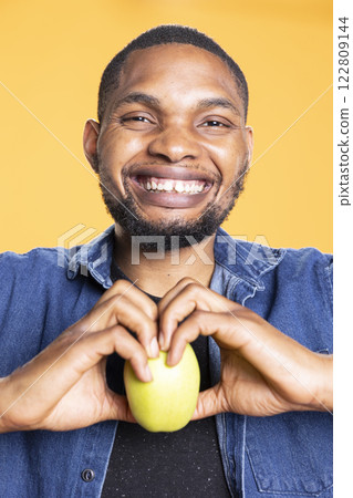 African american smiling adult forms a heart with his hands and holding an apple inside of it, advertising healthy eating zero waste. Vegan cheerful man recommending natural organic goods. 122809144