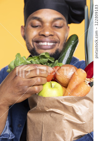 African american chef admiring organic produce in a paper bag, smelling the fresh aroma of fruits and vegetables in studio. Person cooking with bio natural ingredients. Close up. African american chef admiring organic produce in a paper bag, smelling the fresh aroma of fruits and vegetables in studio. Person cooking with bio natural ingredients. Close up. 122809183