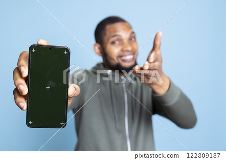 Portrait of african american guy showing chroma key screen on smartphone, creating a commercial ad against blue background. Young person smiling and recommending something in studio. Portrait of african american guy showing chroma key screen on smartphone, creating a commercial ad against blue background. Young person smiling and recommending something in studio. 122809187