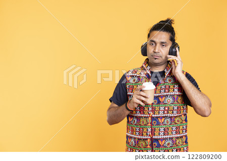 Tired indian man listening music, enjoying hot beverage. Person wearing headphones, hearing songs, drinking coffee to wake up and gain energy, isolated over studio background Tired indian man listening music, enjoying hot beverage. Person wearing headphones, hearing songs, drinking coffee to wake up and gain energy, isolated over studio background 122809200