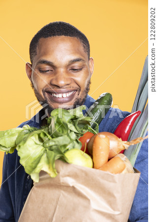 Smiling cheerful adult smiling at his fresh organic produce, feeling happy with his grocery store shopping session. Vegan young guy carrying his paper bag full of ethically sourced bulk products. 122809202