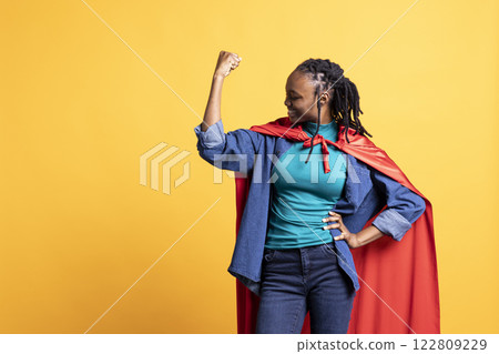 Happy BIPOC girl portraying superhero with red cape flexing muscles, isolated over studio background. Cheerful young woman wearing cloak posing as hero in costume, having fun Happy BIPOC girl portraying superhero with red cape flexing muscles, isolated over studio background. Cheerful young woman wearing cloak posing as hero in costume, having fun 122809229