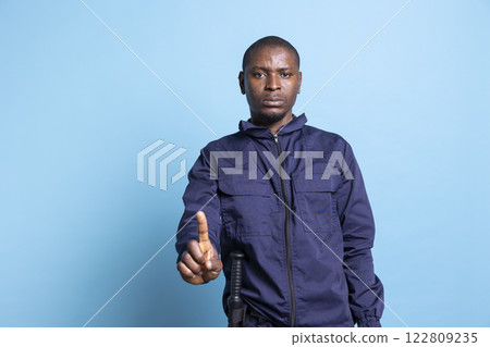 African american security agent creates a stop symbol in front of the camera, restricting the access with a symbol against blue background. Man working as a bodyguard forbidding something. 122809235