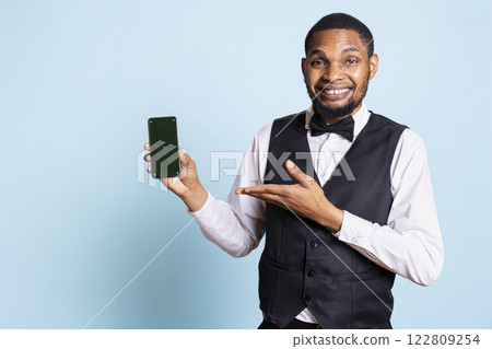 Hotel concierge staff holding a smartphone with chroma key display, showing copy space against blue background in studio. Hotelier bellhop welcoming resort guests at reception. 122809254