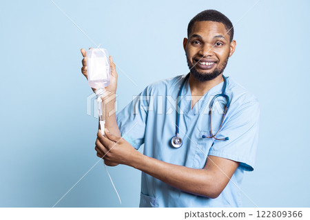 Male nurse holding IV bag, preparing to use it to administer fluids, medications and nutrients directly into patient veins, studio background. Man holding intravenous therapy medical tool. Male nurse holding IV bag, preparing to use it to administer fluids, medications and nutrients directly into patient veins, studio background. Man holding intravenous therapy medical tool. 122809366