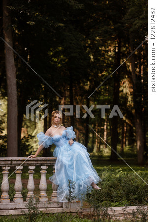 Romantic Woman in a Light Blue Gown Posing on a stone balustrade in a picturesque garden. Romantic Woman in a Light Blue Gown Posing on a stone balustrade in a picturesque garden. 122812432