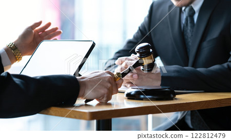 Business professionals in formal black suits discussing legal matters at wooden desk with judge's gavel, tablet, and documents, for law, justice, or corporate negotiation 122812479