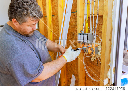 An electrician works installing insulation spray foam on electrical connections in wall framing during home renovations. 122813058