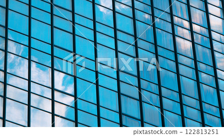 a reflective glass facade with a grid-like design, showcasing the sky and clouds mirrored on the surface. It creates a dynamic interplay of light, color, and symmetry. 122813251