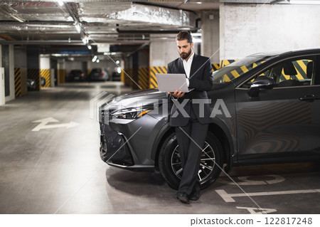 Caucasian male businessman using laptop while leaning on car in underground parking garage, showcasing modern work lifestyle and technology. Caucasian male businessman using laptop while leaning on car in underground parking garage, showcasing modern work lifestyle and technology. 122817248