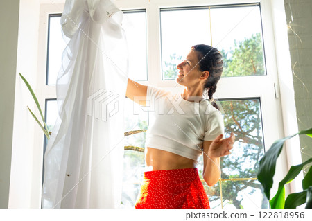 Woman hangs transparent tulle curtains on large windows in the house inside the interior with potted plants. Spring cleaning, tidying up Woman hangs transparent tulle curtains on large windows in the house inside the interior with potted plants. Spring cleaning, tidying up 122818956