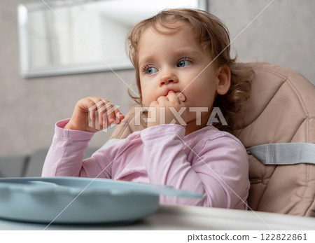 A thoughtful toddler focused on self-feeding while chewing a piece of sausage. A thoughtful toddler focused on self-feeding while chewing a piece of sausage. 122822861