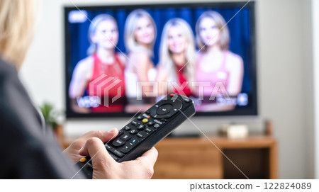 Person holding TV remote control, watching television with blurred faces of women on screen, cozy living room interior, wooden furniture, focus on hand and remote Person holding TV remote control, watching television with blurred faces of women on screen, cozy living room interior, wooden furniture, focus on hand and remote 122824089