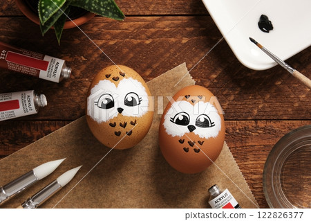 Easter eggs with owls, hand-painted with acrylic paints, on a background of paints, brushes, and palette. DIY concept 122826377