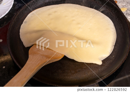 A wooden spatula lifts a semi-cooked, golden-brown crepe from a well-used black frying pan, capturing the skill and precision of home cooking. 122826592