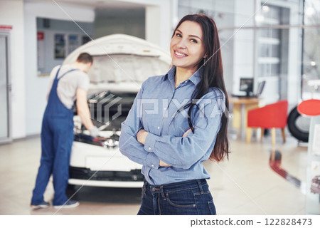 Woman at a car garage getting mechanical service. The mechanic works under the hood of the car Woman at a car garage getting mechanical service. The mechanic works under the hood of the car 122828473