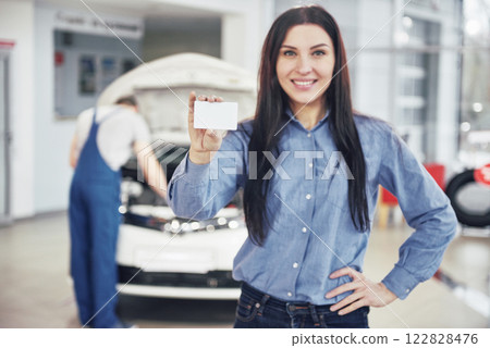 A beautiful woman keeps a business card of the car service center. The mechanic inspects the car under the hood in the background A beautiful woman keeps a business card of the car service center. The mechanic inspects the car under the hood in the background 122828476