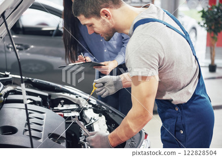 A man mechanic and woman customer look at the car hood and discuss repairs A man mechanic and woman customer look at the car hood and discuss repairs 122828504