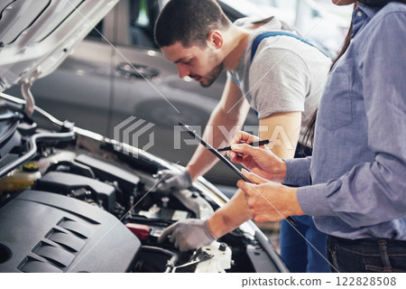 A man mechanic and woman customer look at the car hood and discuss repairs A man mechanic and woman customer look at the car hood and discuss repairs 122828508