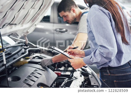 A man mechanic and woman customer look at the car hood and discuss repairs A man mechanic and woman customer look at the car hood and discuss repairs 122828509
