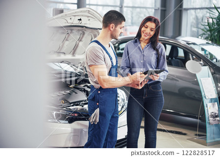 Beautiful young woman is talking to handsome car mechanic while repair a car in dealership Beautiful young woman is talking to handsome car mechanic while repair a car in dealership 122828517