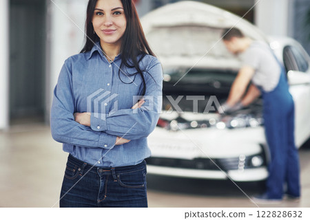 Woman at a car garage getting mechanical service. The mechanic works under the hood of the car 122828632