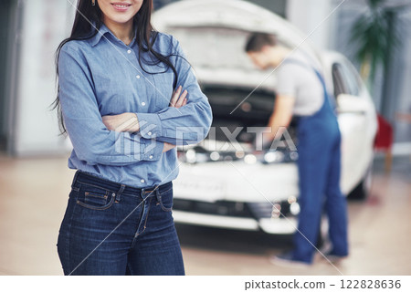Woman at a car garage getting mechanical service. The mechanic works under the hood of the car 122828636